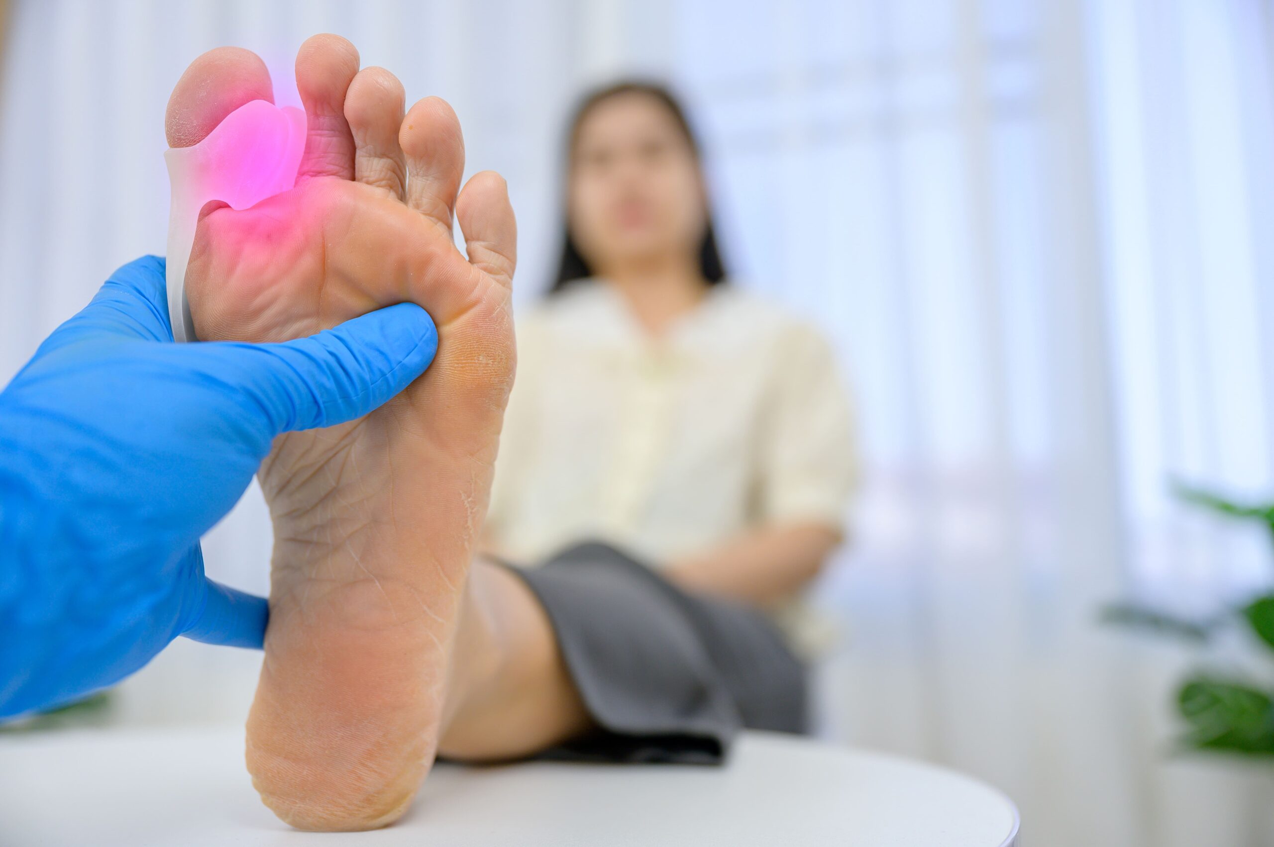 A healthcare professional wearing blue gloves examines a patient's foot, focusing on a red inflamed area around the hallux valgus.